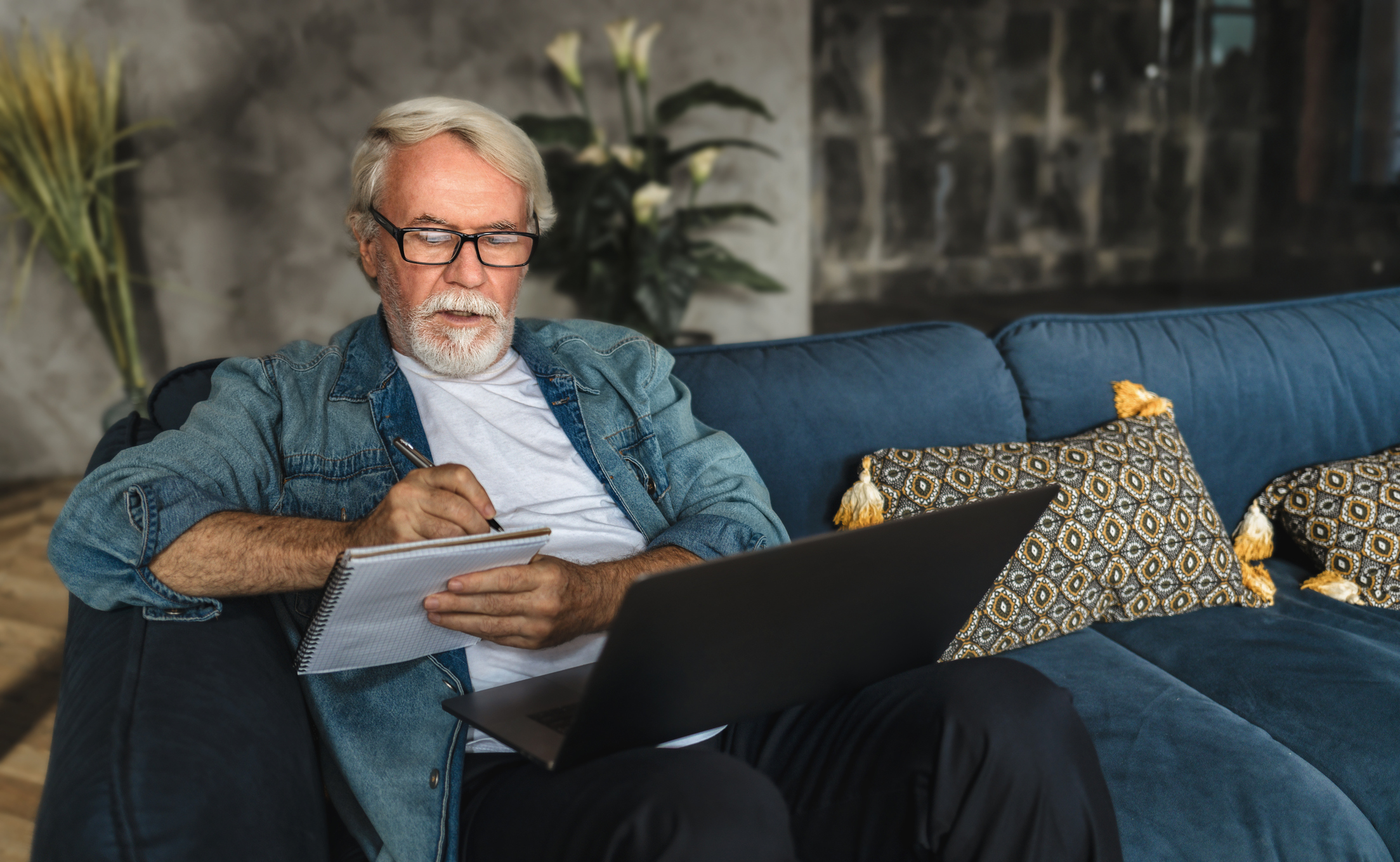 Serious person holding laptop and writing note in notebook.