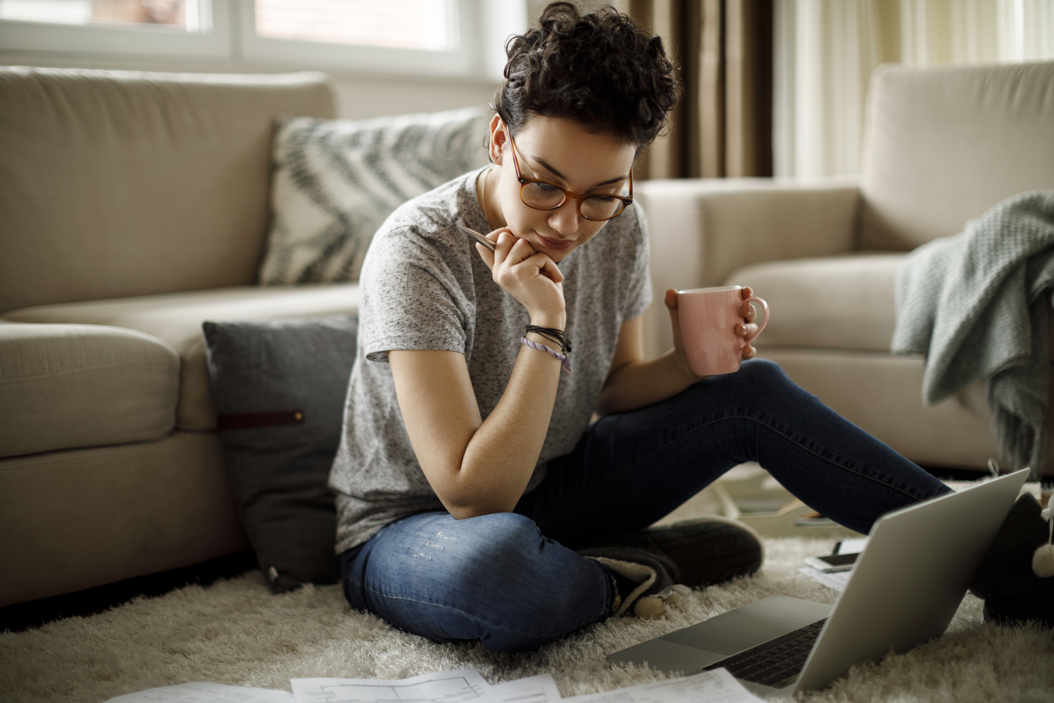 A person holding a mug while sitting on a living room floor and looking at paperwork.