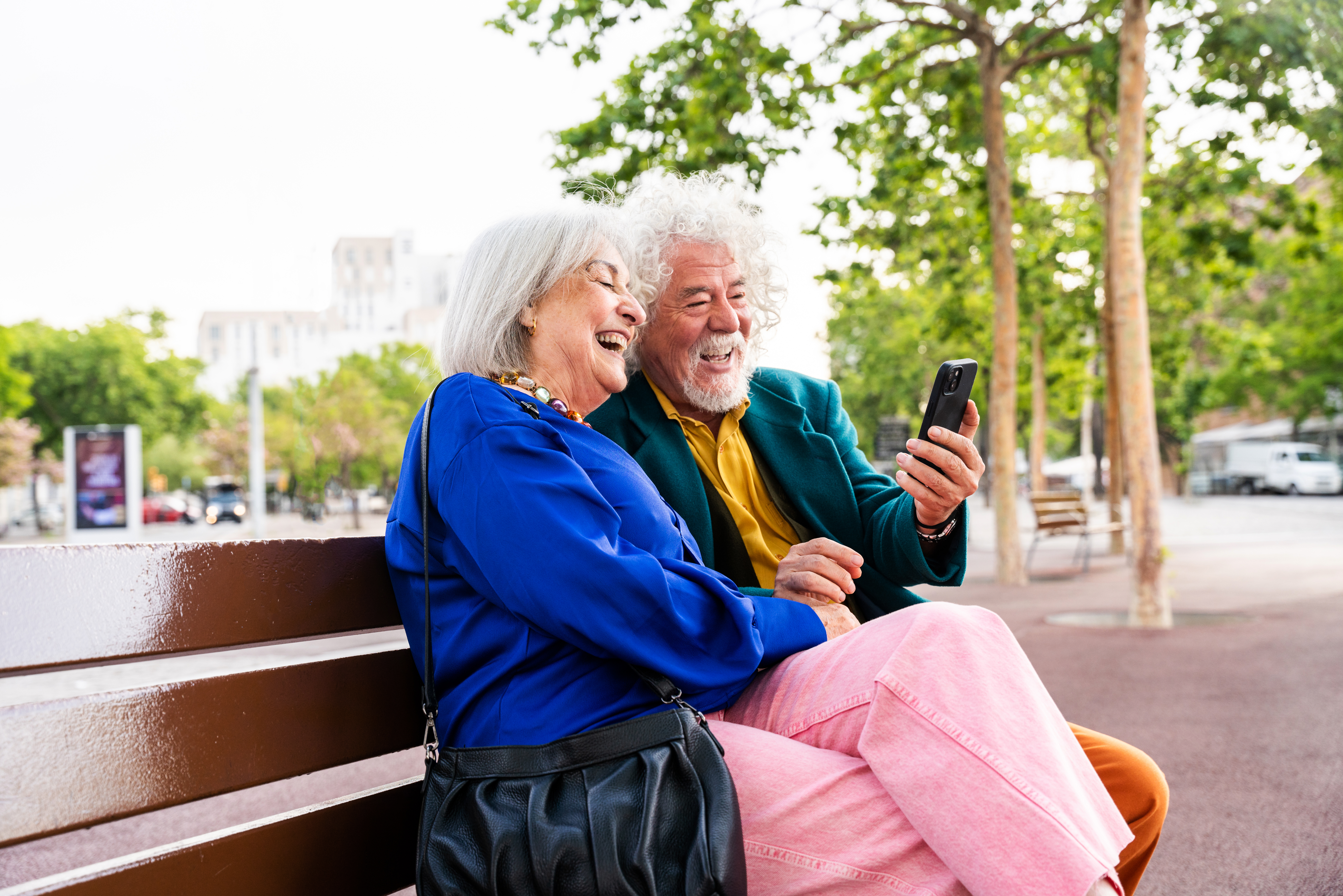 Two smiling people on a bench looking at a phone.