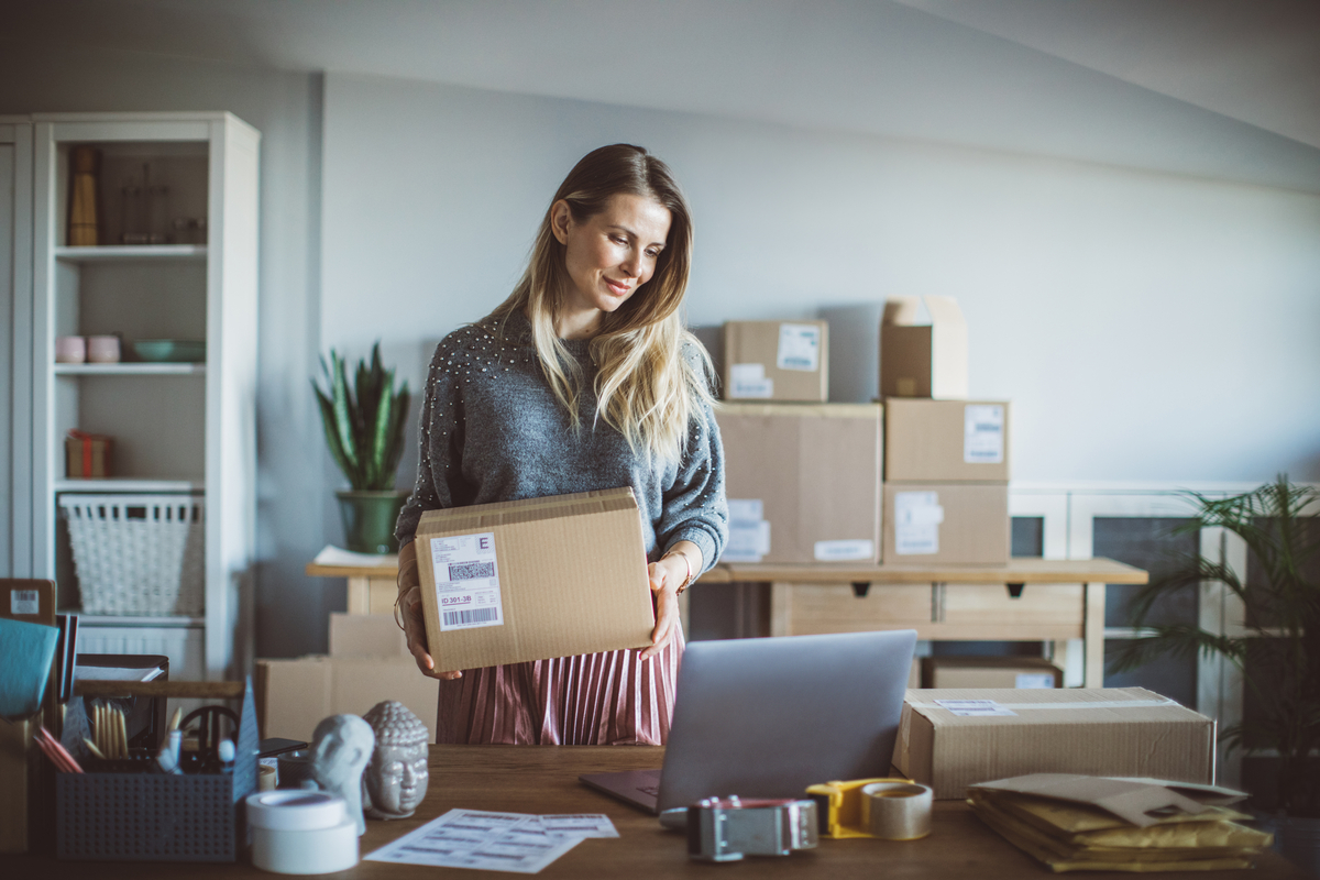 Someone standing and holding a shipping box while looking down toward a laptop.