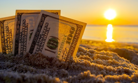 Three fanned one hundred dollar bills partially buried and stood upright in sand, with the sun rising in the horizon.