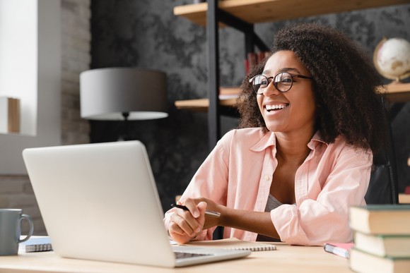 A person smiles while holding a pen and sitting at a table with a notepad and a laptop computer. 