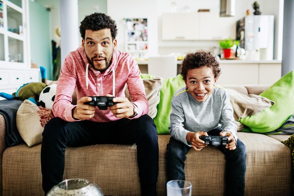 A parent and child holding video game controllers in their hands while seated next to each other on a couch.