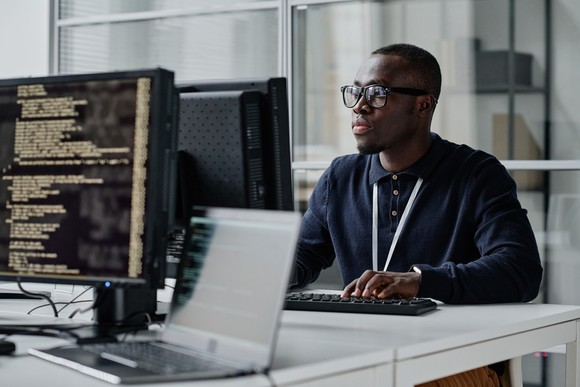 A person sitting at a table working on a computer with other monitors displaying code.  