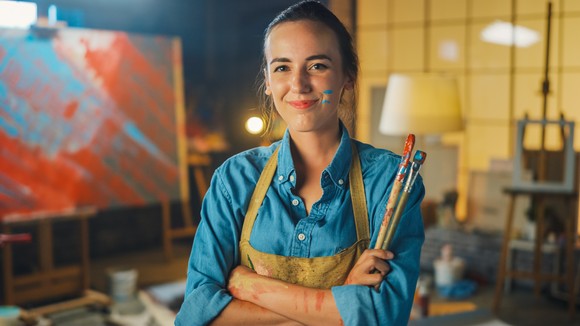 Someone in apron standing in front of painting, holding paintbrushes and smiling. 