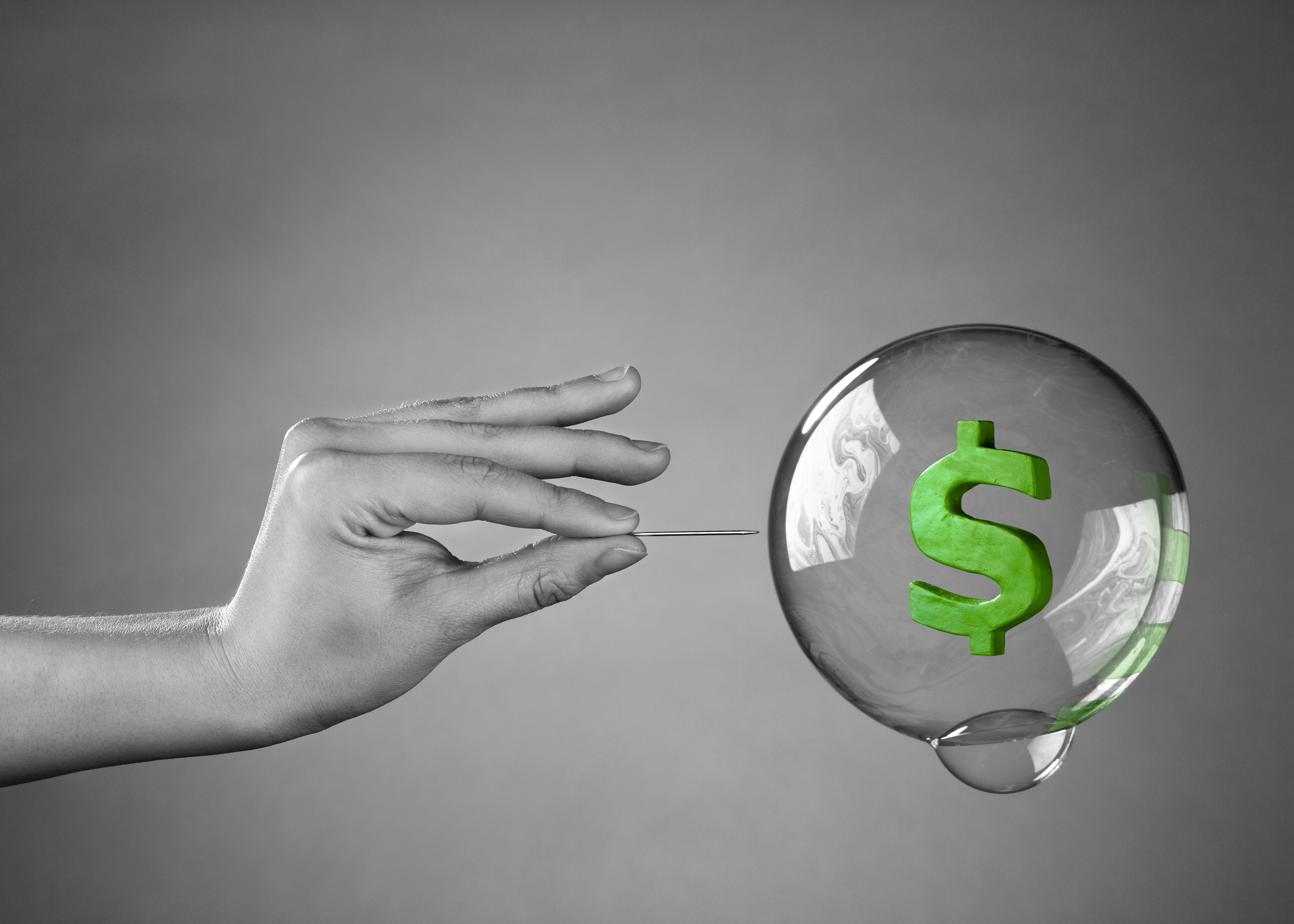 A hand holding a pin that's being used to to pop a bubble containing a green dollar sign.