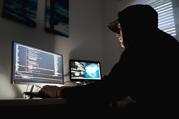 A hacker wearing a concealing hoodie who's typing on a keyboard in a dimly-lit room. 