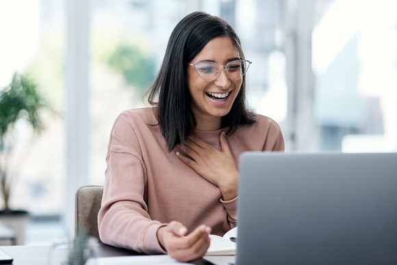 A person smiling while sitting at a table in-front of a laptop computer.