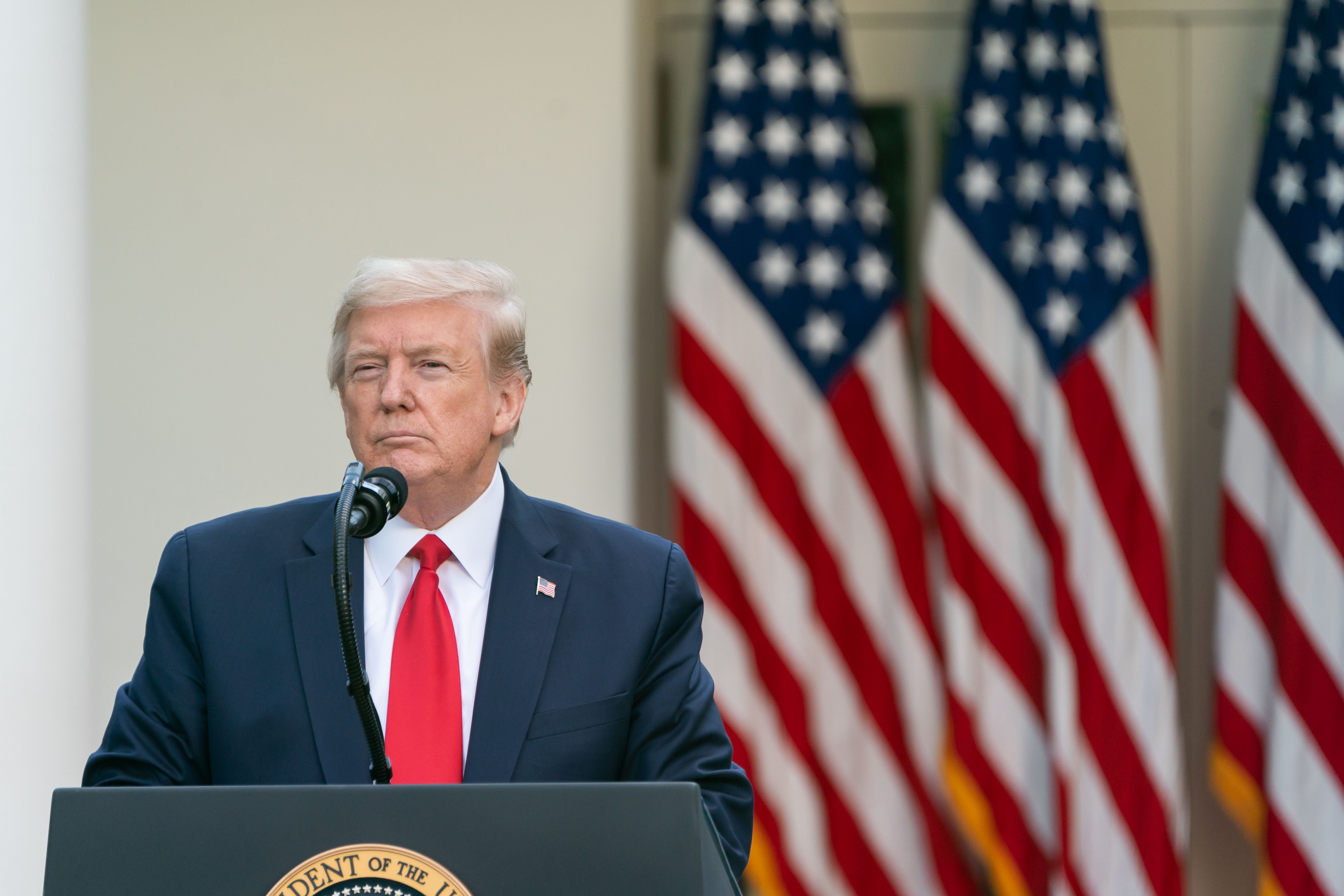 Donald Trump stands at a podium with American flags displayed in the background.