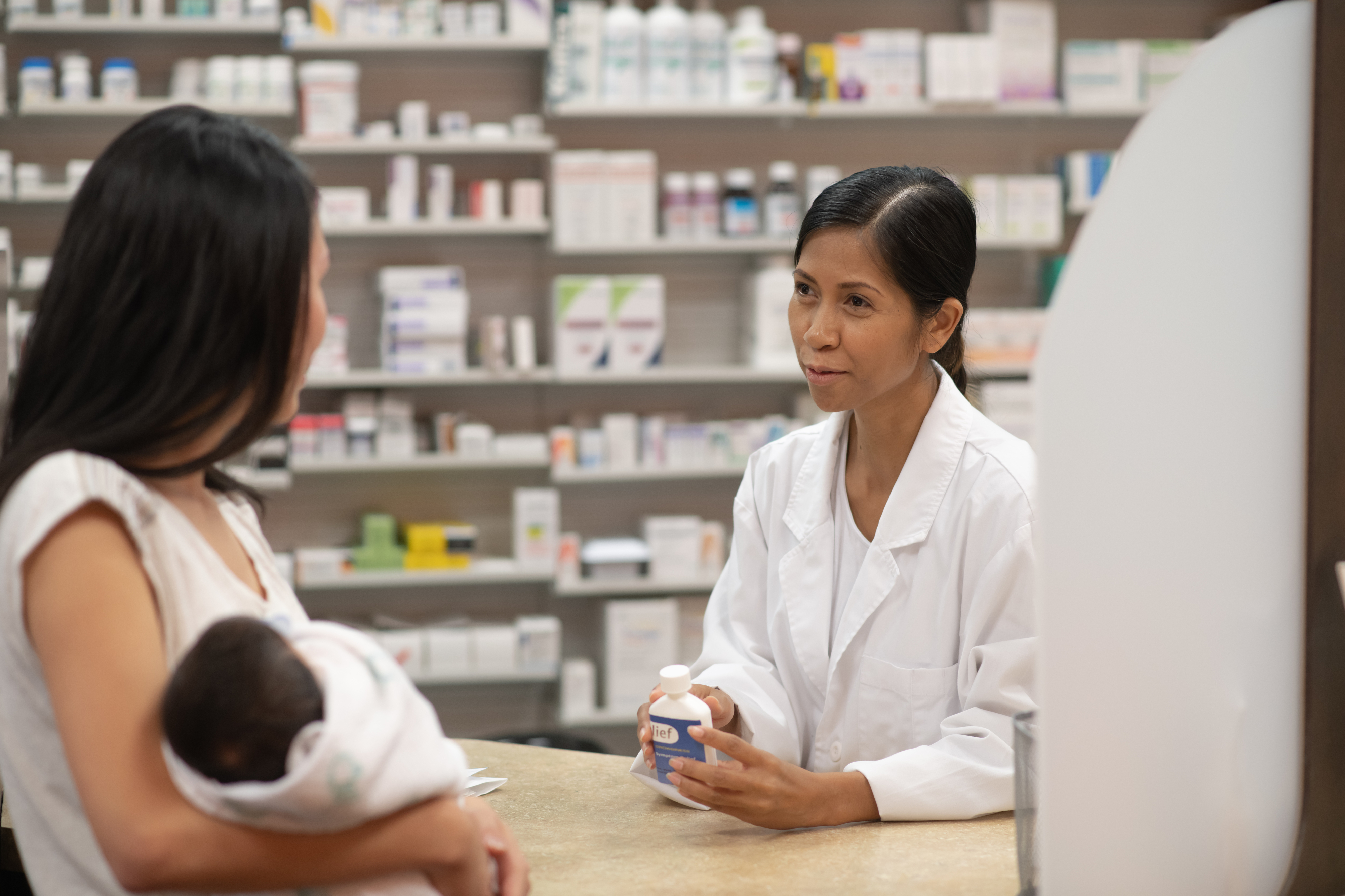 A pharmacist holding a prescription while speaking with a customer who has an infant in their arms.