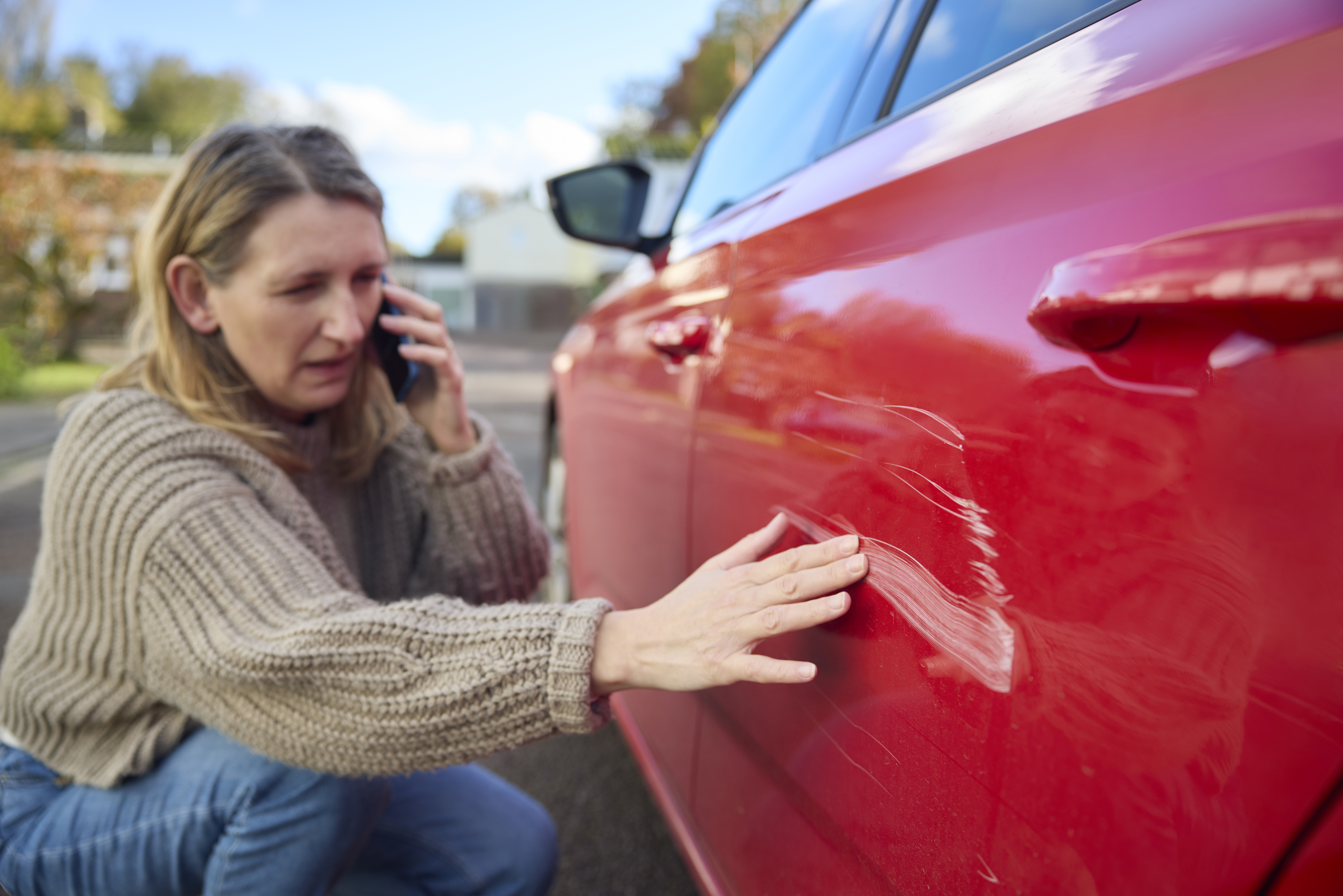 A person is on the phone while looking at a scratch on their car.