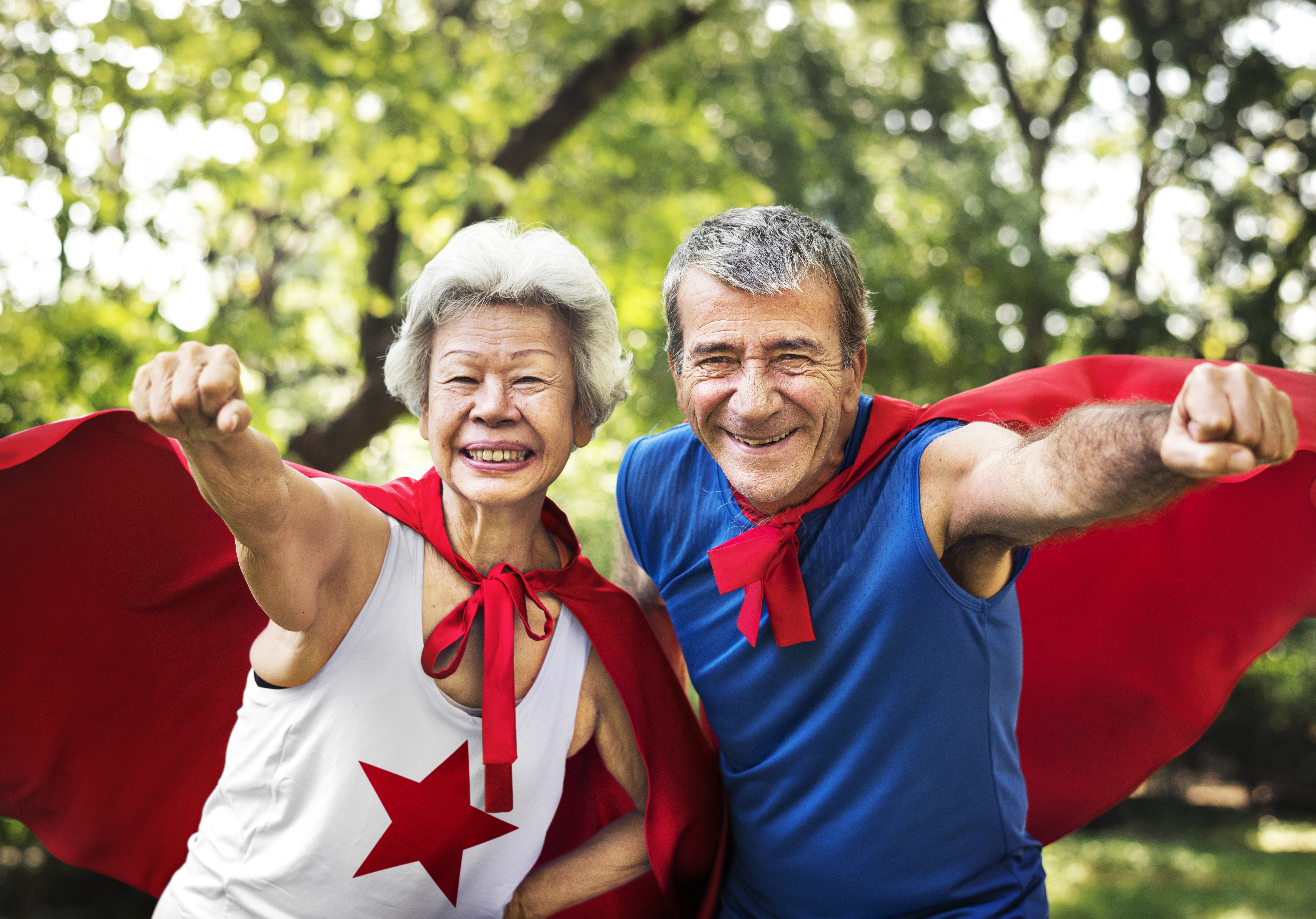 A couple is wearing superhero tops and capes and smiling, with fists outstretched.