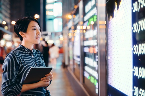 A person holds a tablet and looks at a stock chart and price movements on a city street.