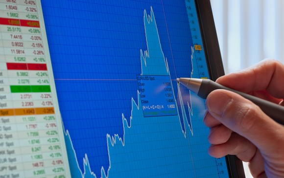 A person's hand holds a pen and traces a stock chart on a computer screen.