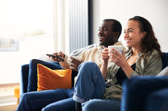 A couple sits on the couch, smiling as they watch television.