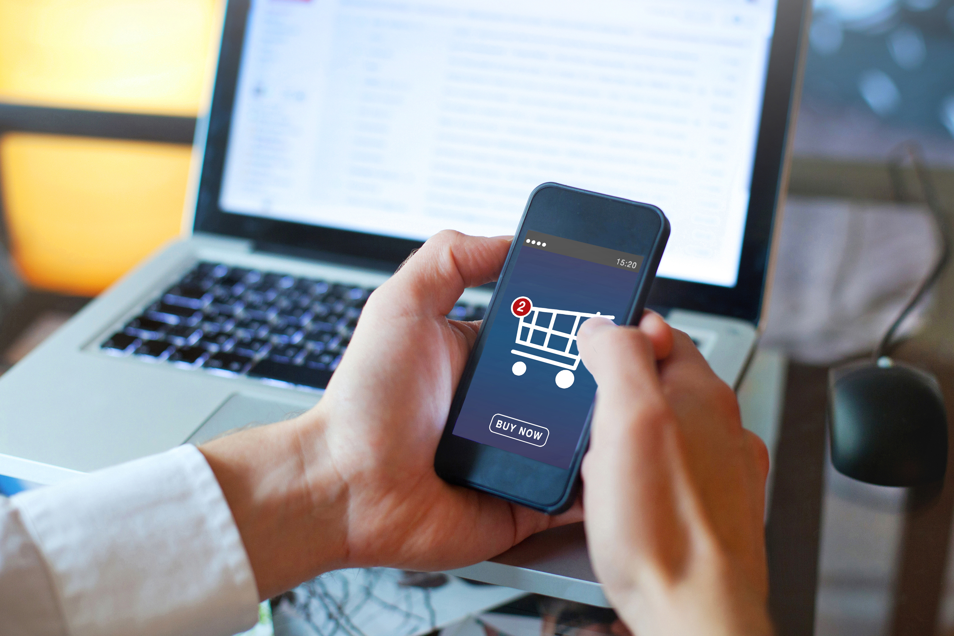 A person shopping online on a mobile phone with a laptop in the background.