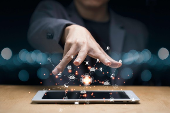 A person wearing a blazer outstretches their arm and opens their hand over a tablet that is emitting various icons, illustrating the concept of technological advancement. 
