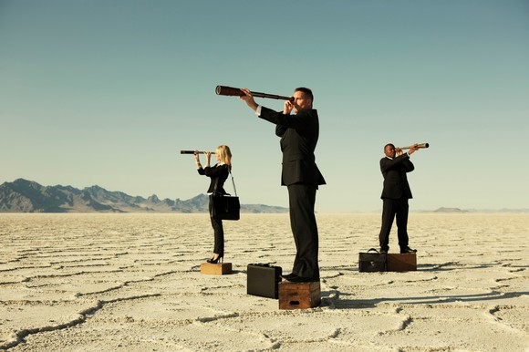 Three people standing on boxes in a desert are looking through telescopes.