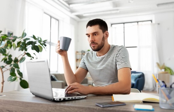 A person sitting at a desk drinking a cup of coffee while using a laptop. 
