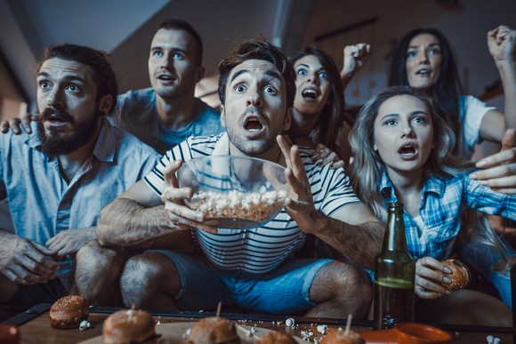 A group of people with popcorn and sandwiches are watching television and looking shocked by what they see. 