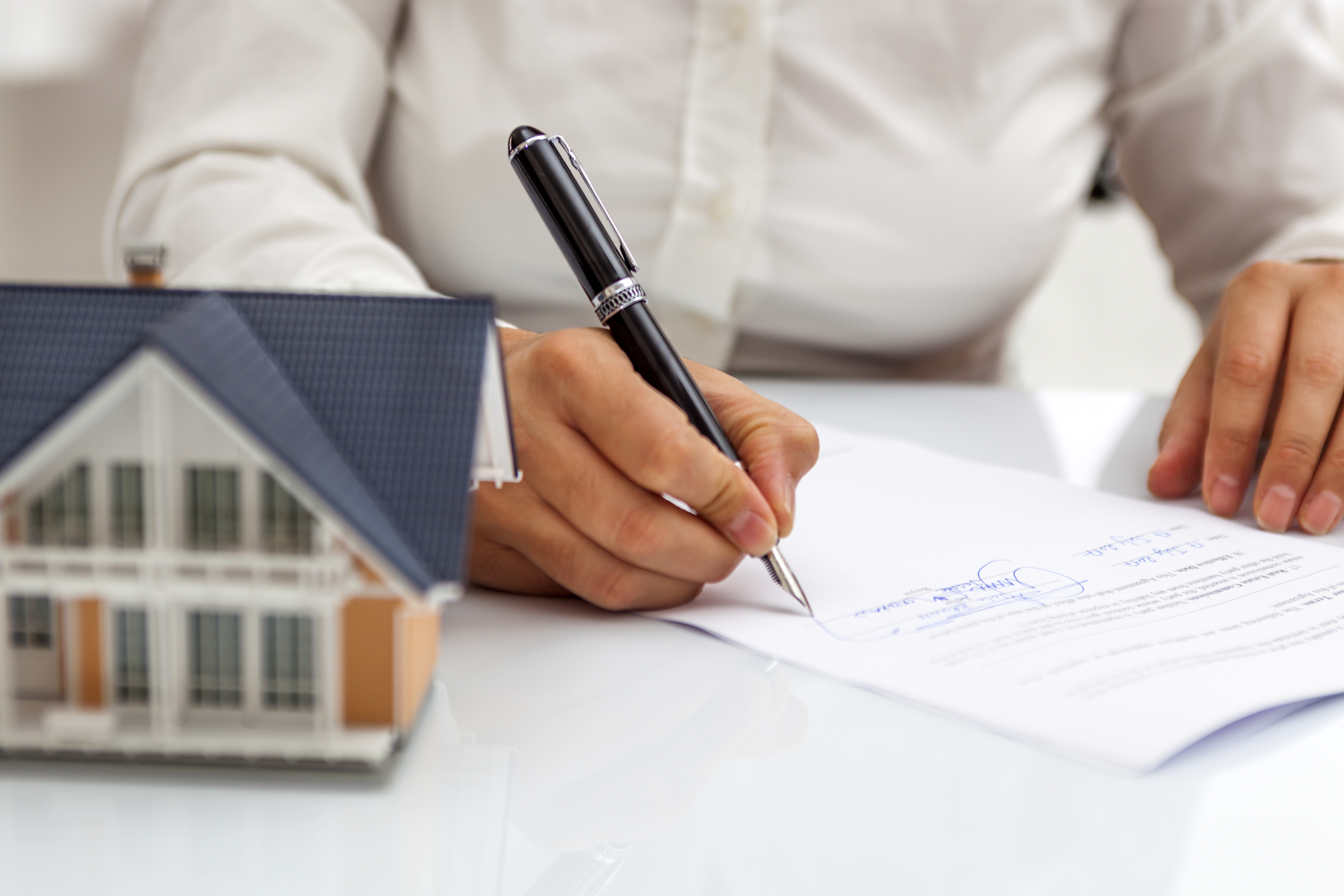 Person signing a document with a small house on the table.