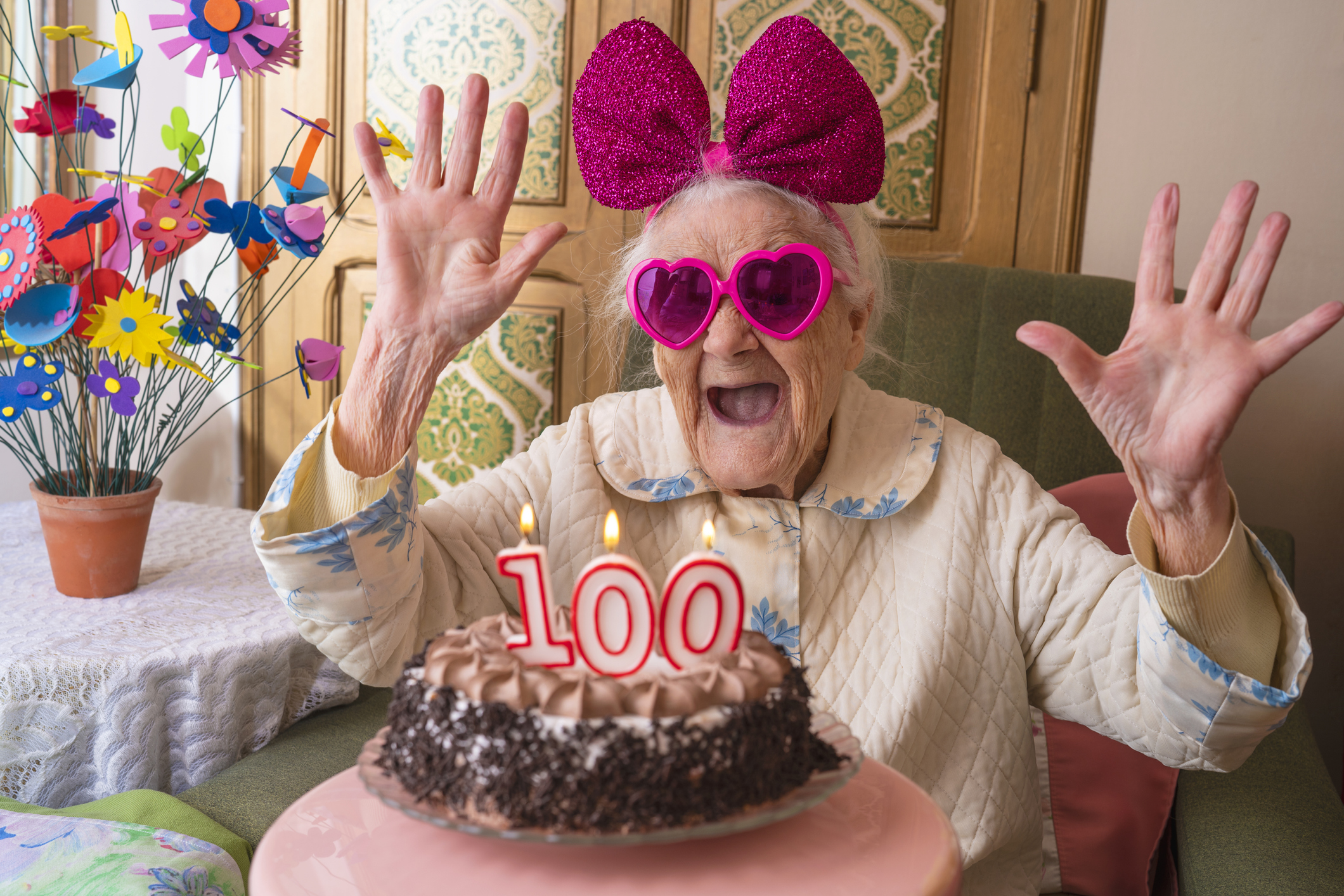 A person smiling and throwing up hands in joy, behind a 100th birthday cake.