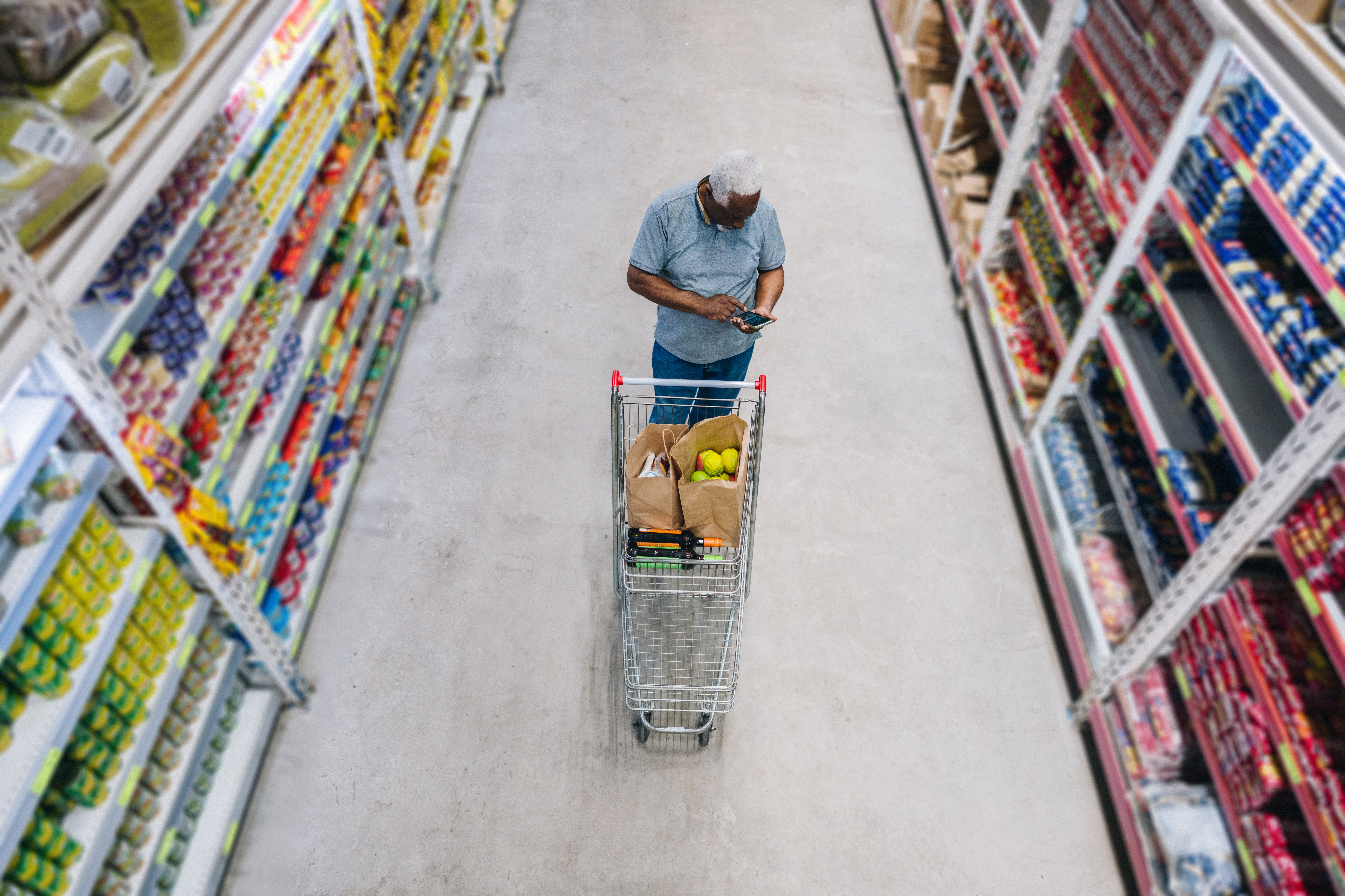 Man looks at phone with shopping cart in empty store aisle. 