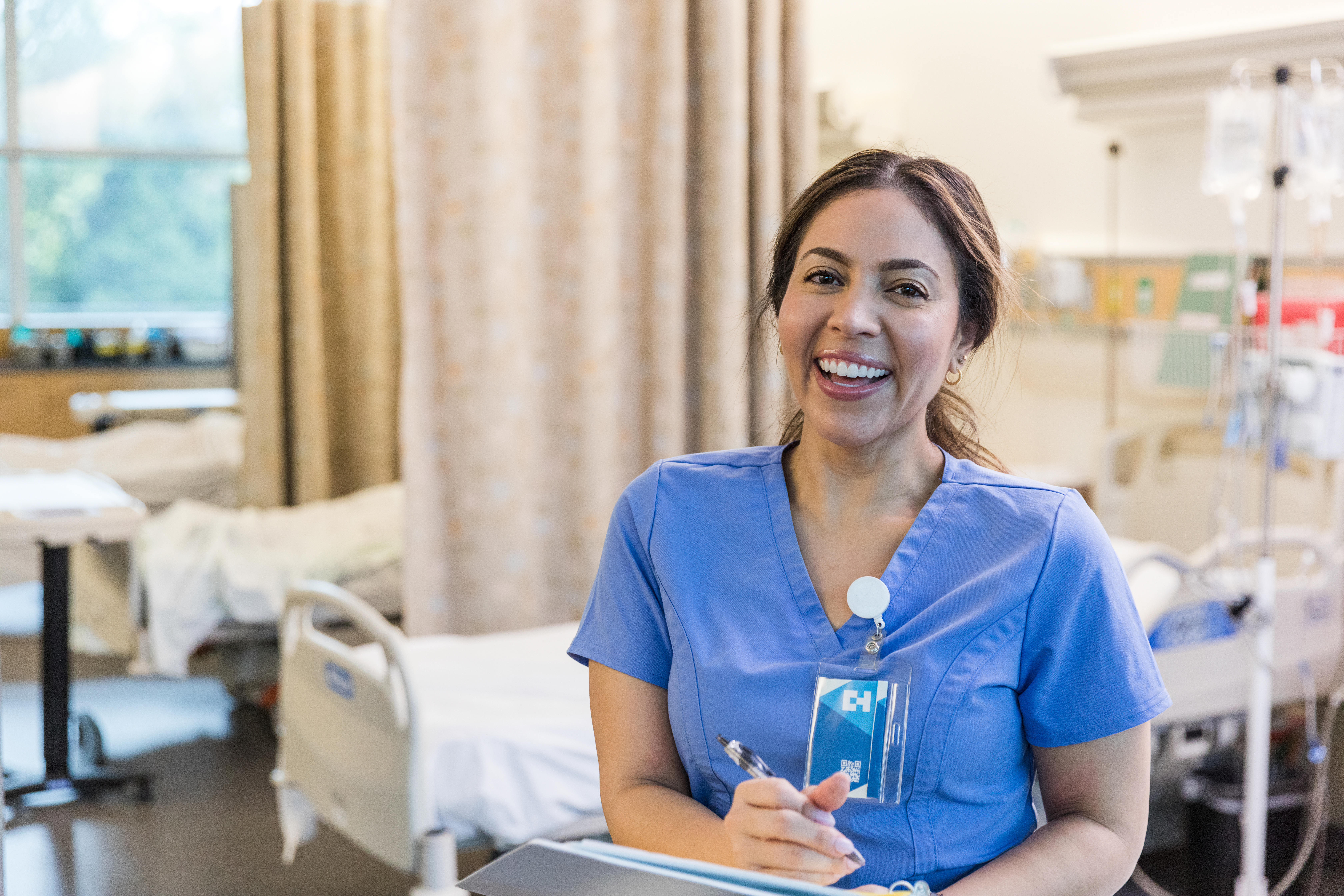 A smiling healthcare worker in blue scrubs is standing in a multiple-bed room in a hospital.