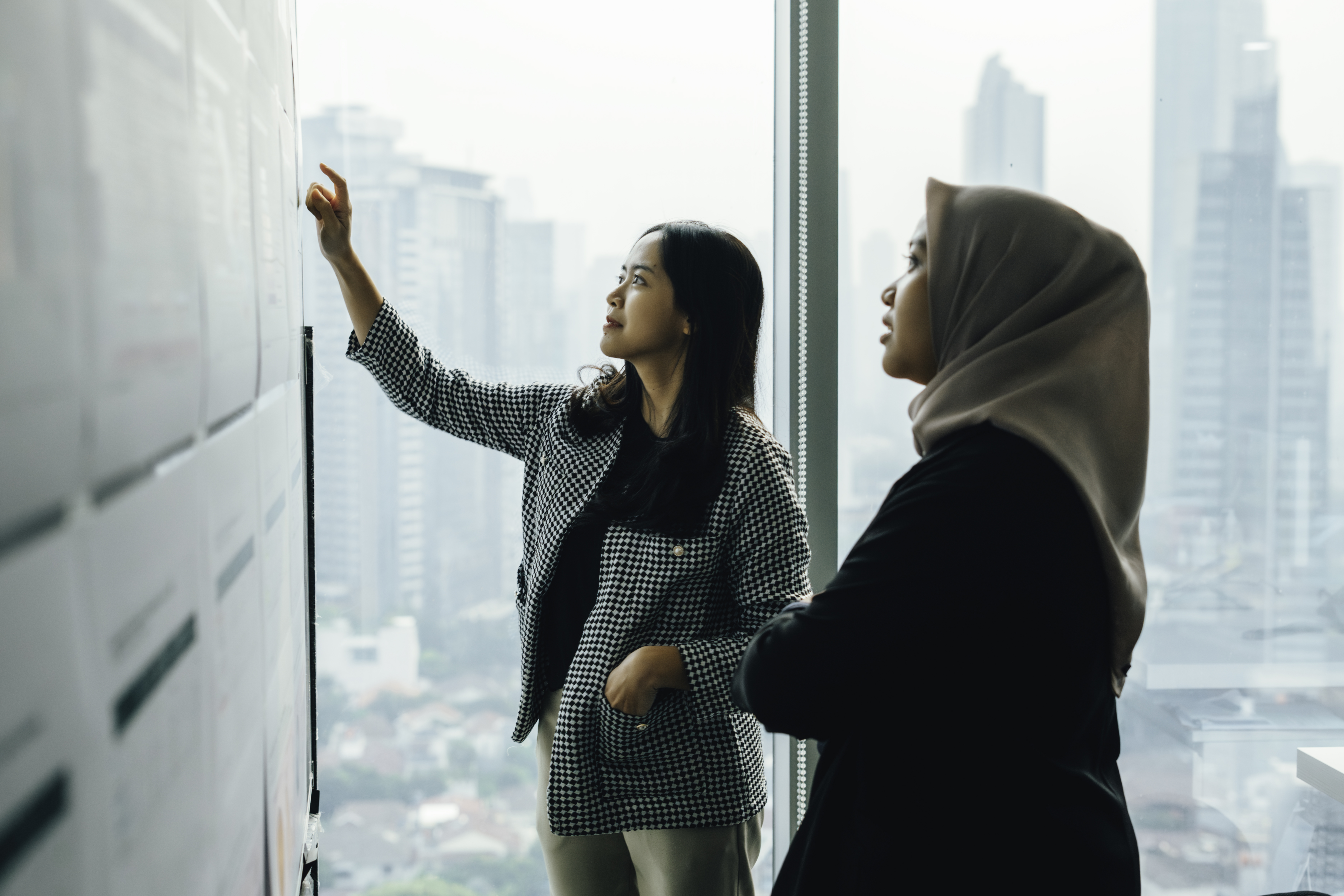 Two people examining papers posted on a wall in an office.