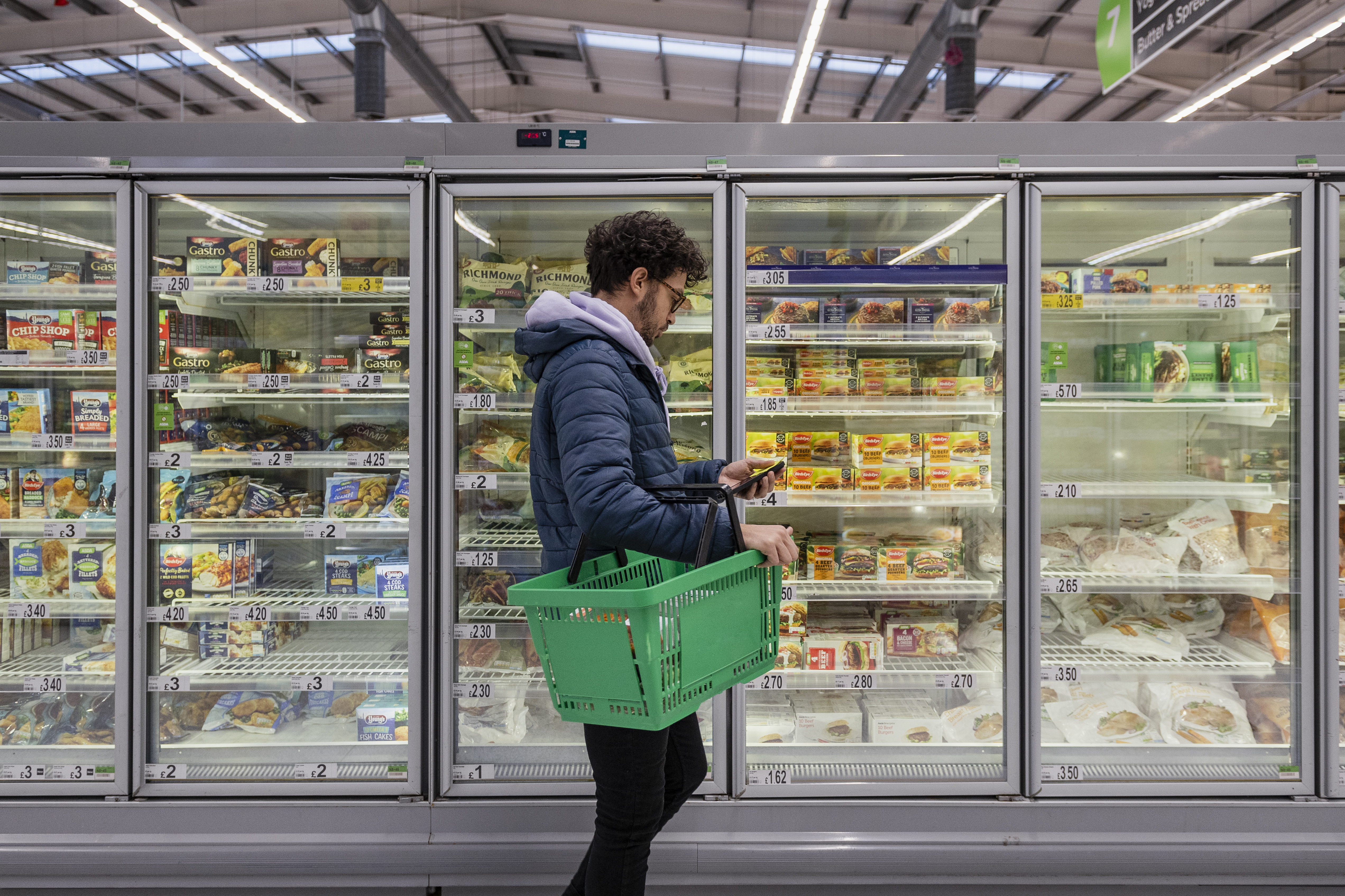 A customer holds a green shopping basket while looking at an item in the frozen foods section of a grocery store.