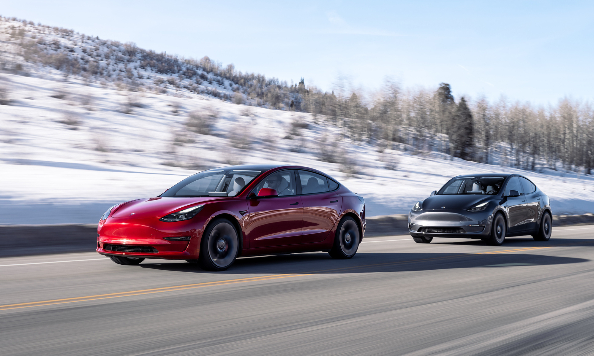 Red and black Tesla EVs driving down road with snow covered hill in background.