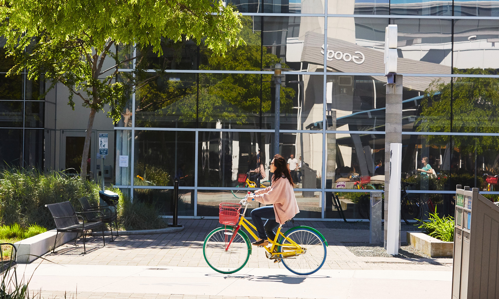 Lady biking on Google campus with Google name reflected on window in background. IS GOOG.
