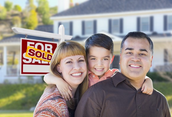 Smiling man, woman, and child in front of house with sold sign