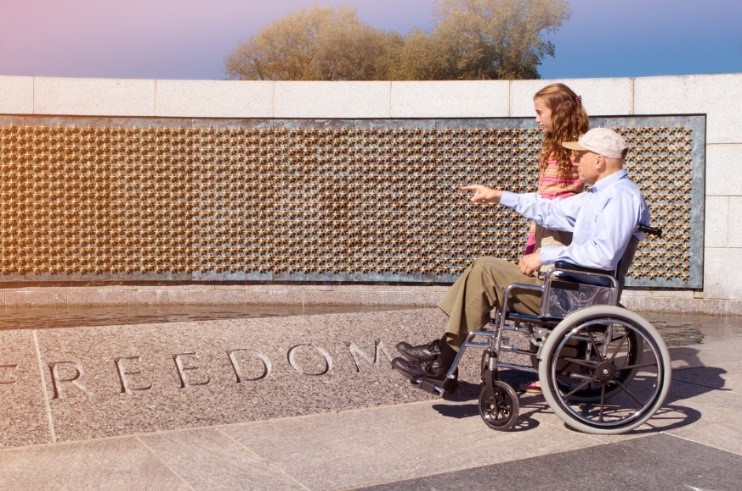 An elderly person using a wheelchair points a companion to a monument reading FREEDOM.