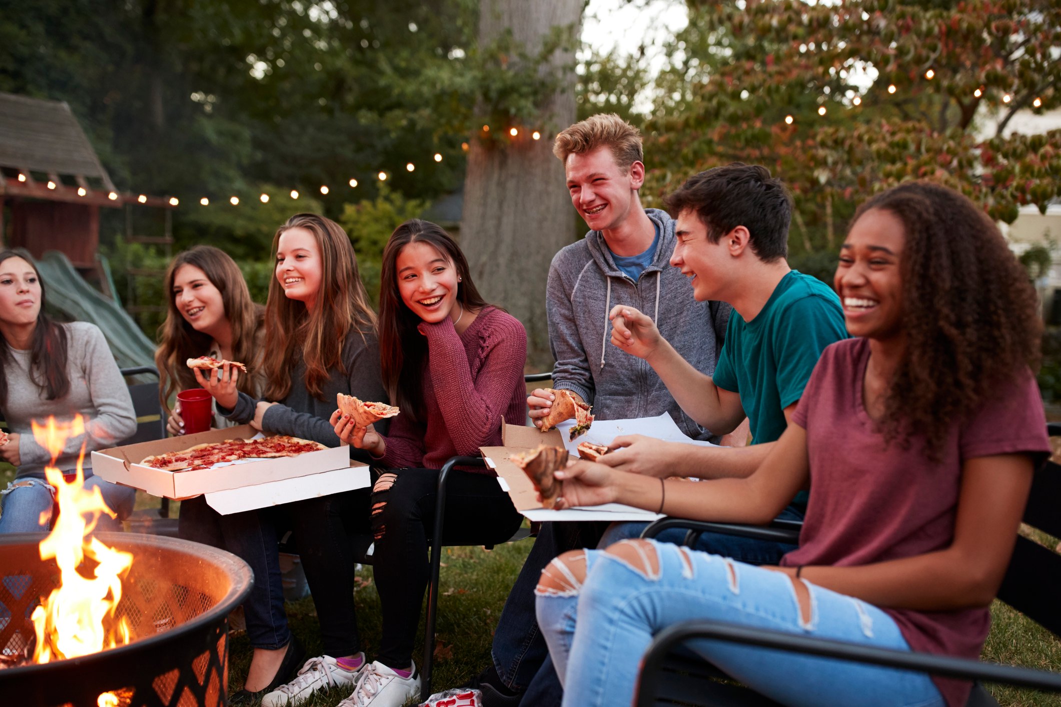 Seven teens gathered around a fire pit while eating delivery pizza