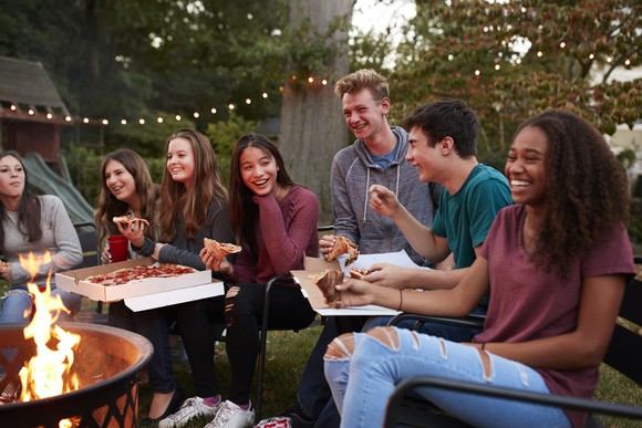 Seven teens gathered around a fire pit while eating delivery pizza