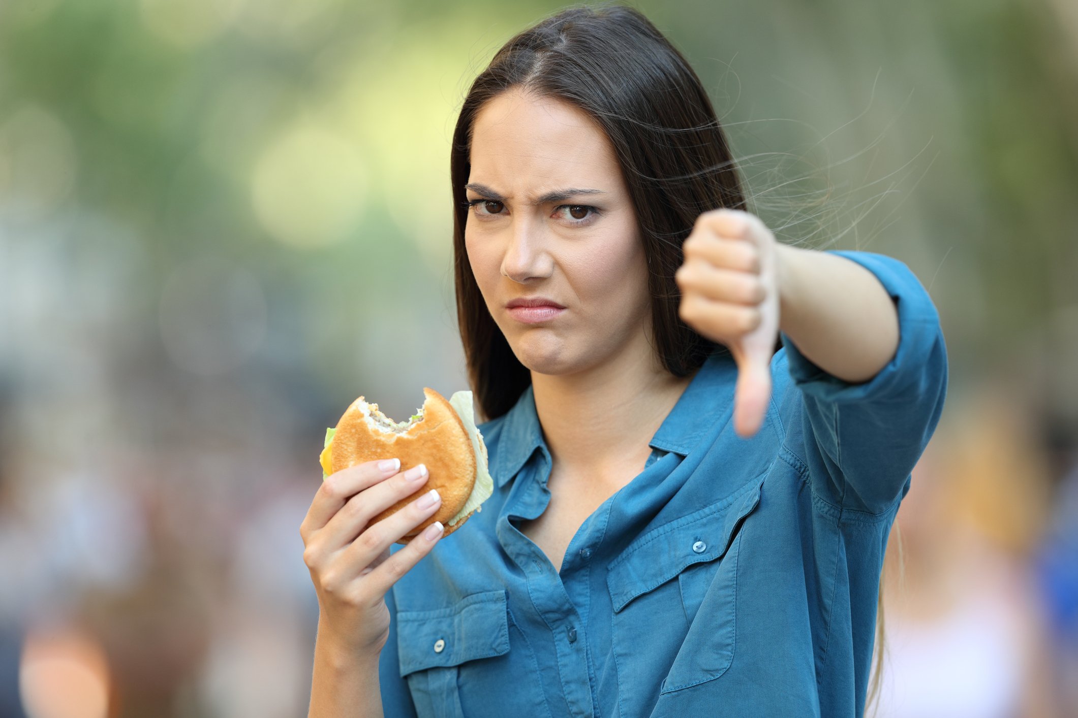 Girl holding a hamburger in one hand and giving a thumbs down with her other hand.
