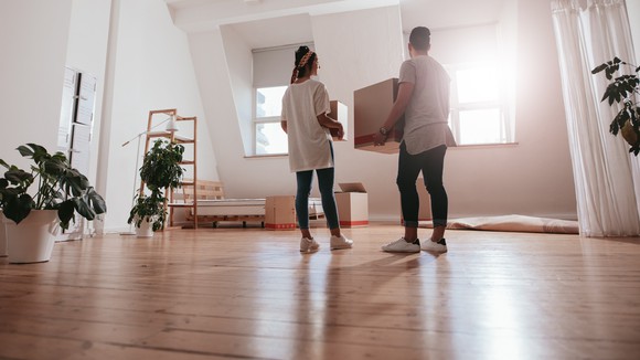 Two people carrying boxes into a mostly empty room in a house