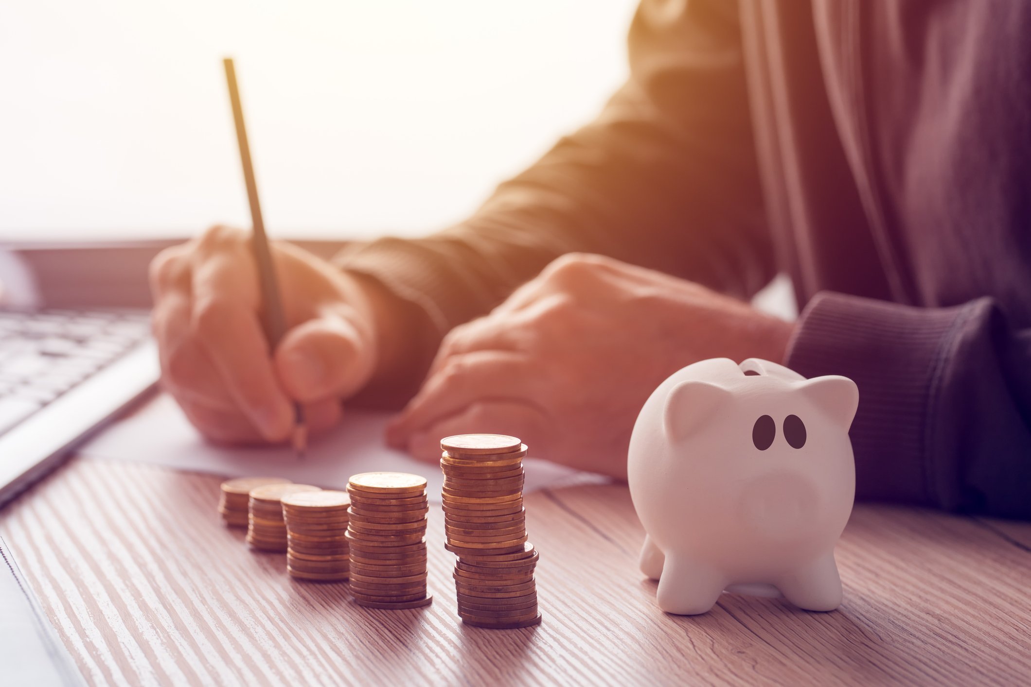 A man writing something with stacks of coins and a piggy bank on the desk next to him