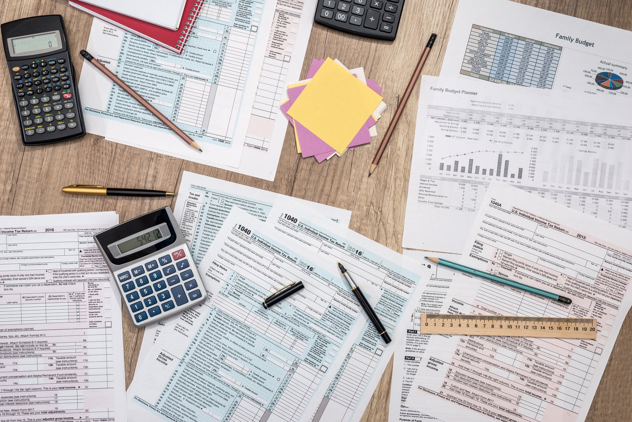 Various tax forms on top of a wooden table, with calculators, pens, a ruler, and Post-its strewn around them.