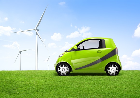 A compact green electric car in a field with windmills in the background.