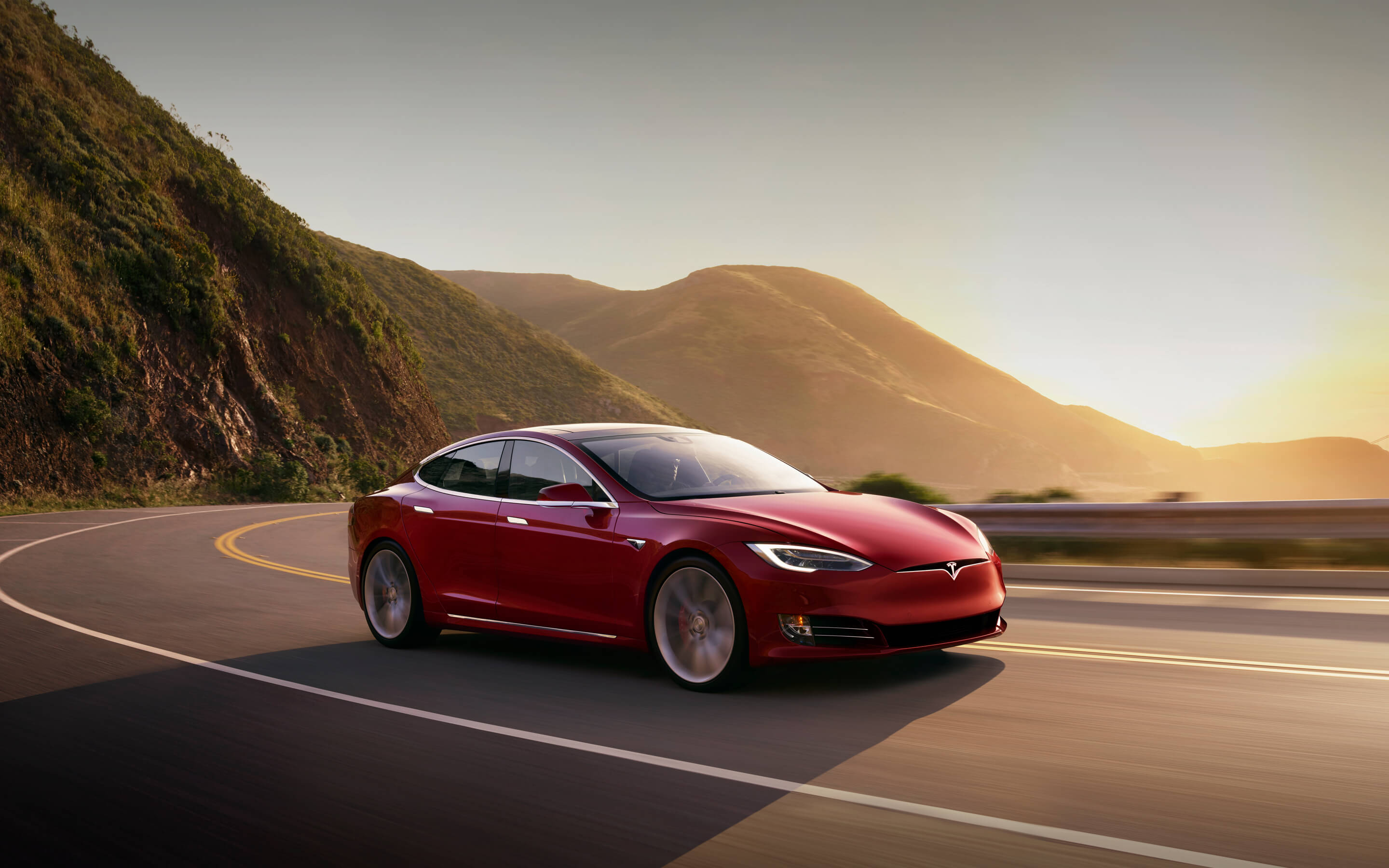 Red Model S driving along curving road with mountains and sun in the background