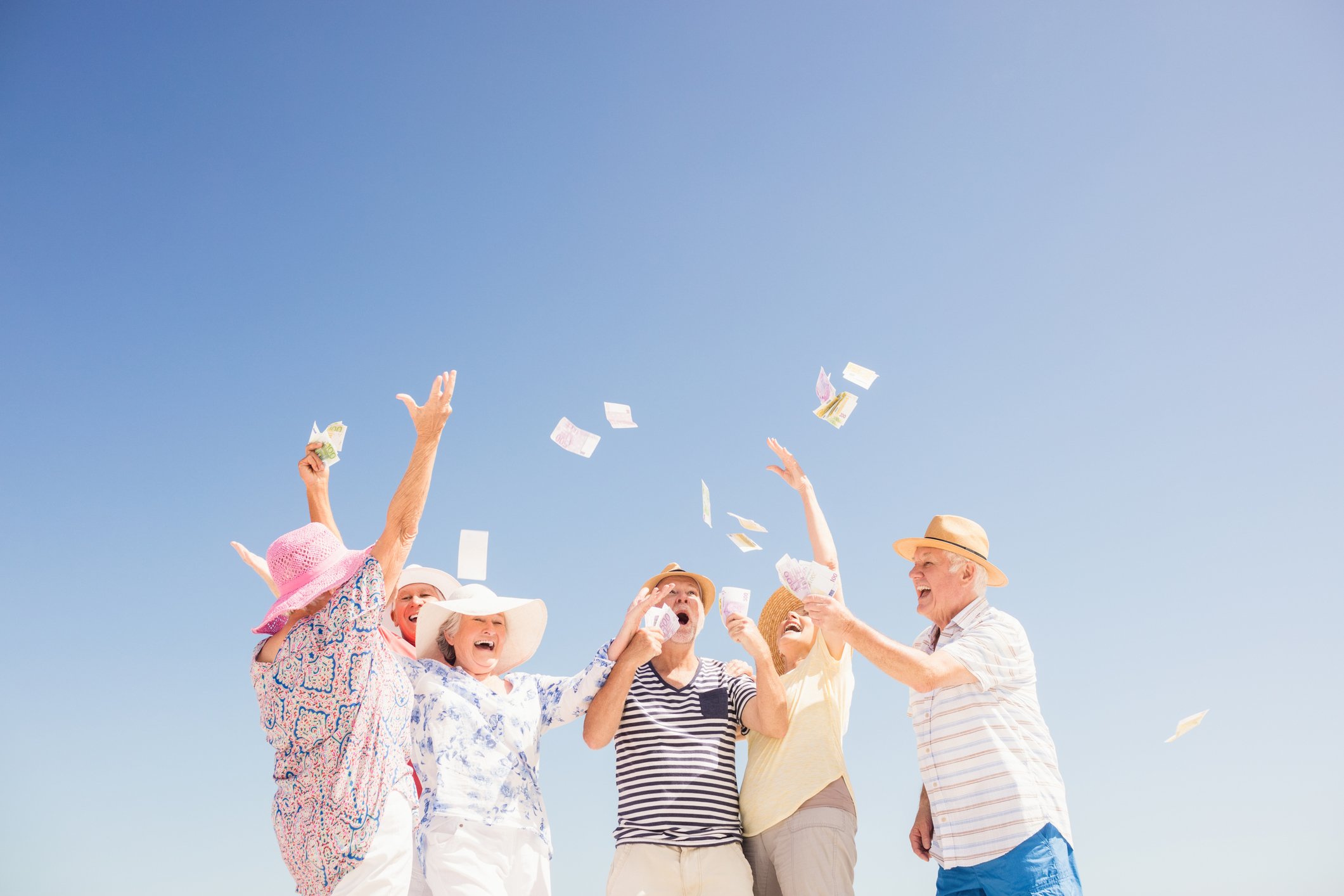 Senior citizens dressed for vacation, happily throwing cash into the air against a blue sky.