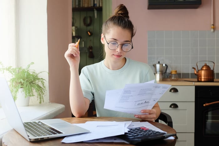 A young woman sits at a kitchen table with papers in hand and a calculator and laptop on the table.