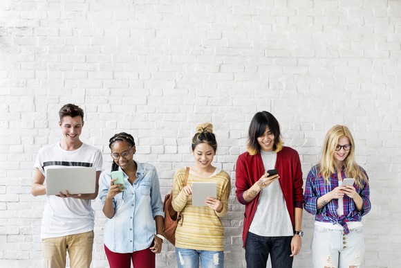 Five young people lined up against a white brick wall while looking at their wireless devices and smiling.