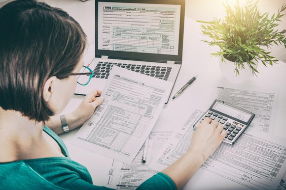A woman calculates taxes on a desk strewn with forms, pens, laptop, and calculator.