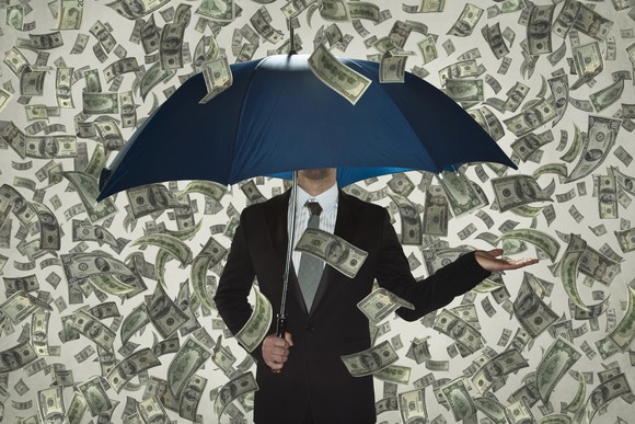 A businessman shelters beneath an umbrella in a shower of hundred-dollar bills.