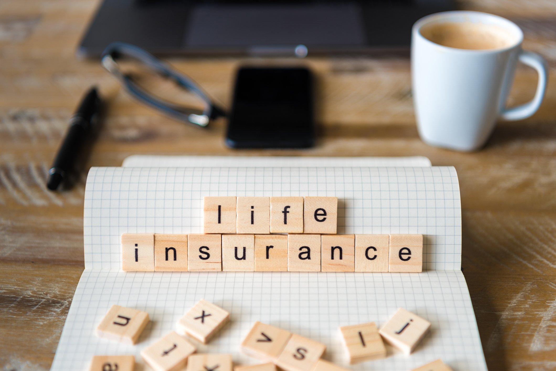 Wooden tiles on a notebook spelling out life insurance with laptop, pen, pair of glasses, smartphone, and coffee cup in the background