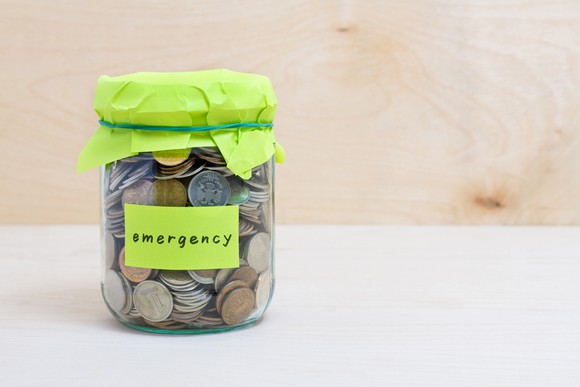 Jar full of coins with a label that says "emergency" sitting on a white tabletop.