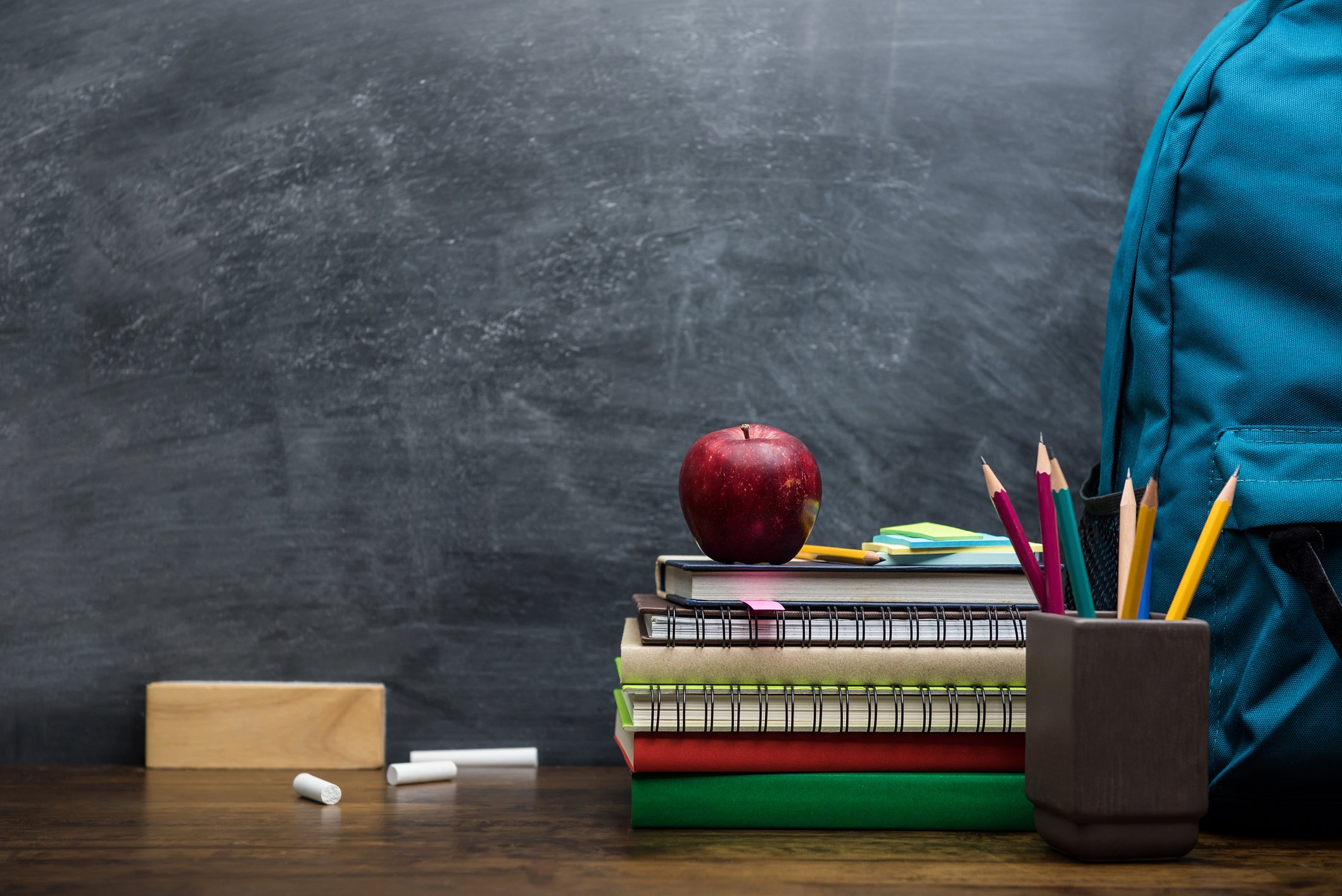 Blackboard, backpack, cup with pencils in it, pieces of chalk, and a stack of books with an apple on top